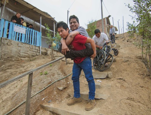 Hombre con discapacidad física siendo apoyado por dos personas para bajar una ladera en un asentamiento humano.